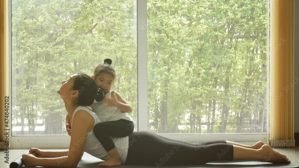4K. Asian mom practice yoga exercise at home with a adorable daughter sitting on mother back. training during quarantine self-isolation. leisure healthy lifestyle. healthy family's activity in the mor 4K. Asian mom practice yoga exercise at home with a adorable daughter sitting on mother back. training during quarantine self-isolation. leisure healthy lifestyle. healthy family's activity in the mor