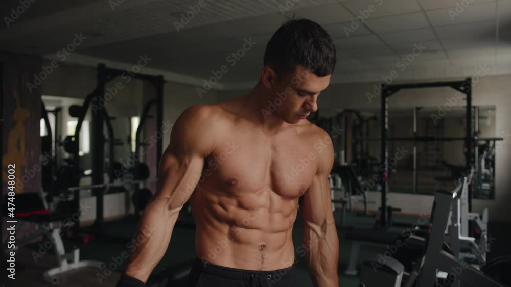 A bodybuilder without a t-shirt shows off his pumped up muscles while standing in the gym and hits his biceps with his fists A bodybuilder without a t-shirt shows off his pumped up muscles while standing in the gym and hits his biceps with his fists