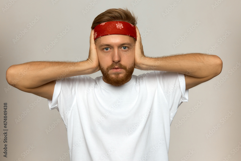 Fototapeta premium Handsome attractive white-skinned man dressed in casual wear and red band over head covering ears avoiding hearing terrible noise, sounds of fighting, feeling lost, looking at camera with anxiety
