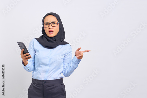 Portrait of surprised young Asian woman holding mobile phone and pointing finger away at copy space isolated on white background photo