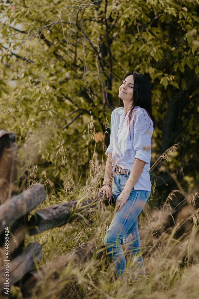 brunette girl with long hair in a white shirt and blue jeans near the fence in the field brunette girl with long hair in a white shirt and blue jeans near the fence in the field
