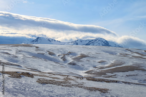 Winter mountain scenery. Snow-capped mountain peaks rise above the snow-covered alpine. In the background is a blue sky and fluffy clouds.