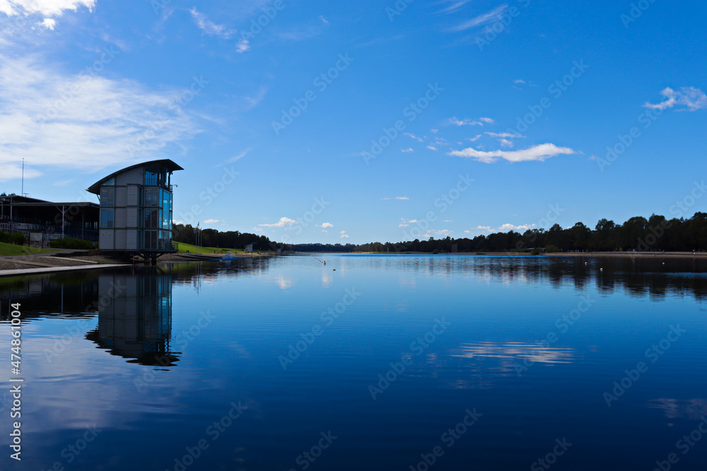 Obraz premium Man made rowing lake with blue sky and white clouds reflecting off almost still water surface. Mountains and trees in the background.