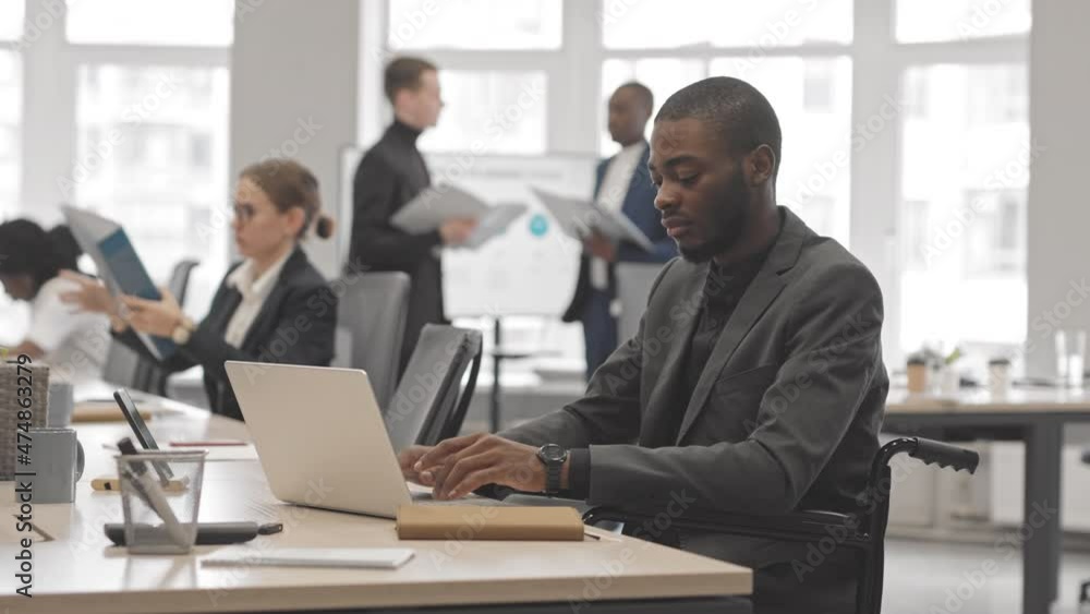 Medium of young Black man in wheelchair sitting at desk in office at daytime, typing on portable computer Medium of young Black man in wheelchair sitting at desk in office at daytime, typing on portable computer