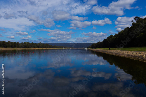 Man made rowing lake with blue sky and white clouds reflecting off almost still water surface. Mountains and trees in the background.