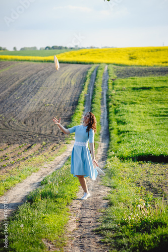 Portrait of stylish girl in hat on green spring background. A young woman in a blue dress walks in nature. Green spring meadow. Girl holding a straw hat over her head.