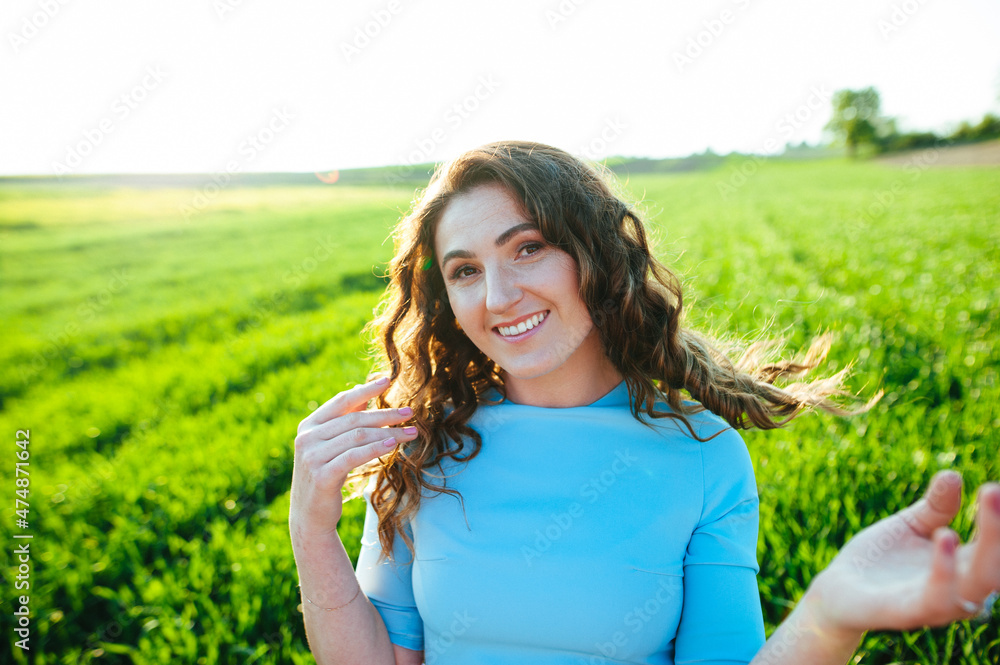 Curly brunette enjoys sunshine in summer day. Portrait of smiling beautiful girl outdoors. Young woman standing on street with curly hair blown by the wind, covering her face. Curly brunette enjoys sunshine in summer day. Portrait of smiling beautiful girl outdoors. Young woman standing on street with curly hair blown by the wind, covering her face.
