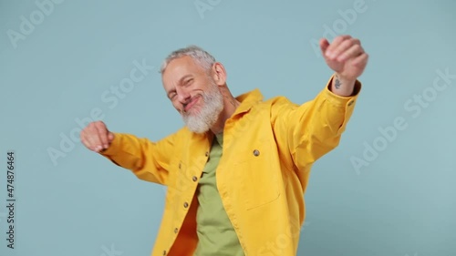 Slow motion fancy vivid elderly gray-haired bearded man 50s in yellow shirt dance fool around have fun gesticulate with hands enjoy relax isolated on plain pastel light blue background studio portrait photo