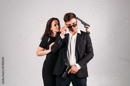 young couple, in black suits with pistols in their hands on a white background in the studio, they depict security guards, bodyguards or agents.