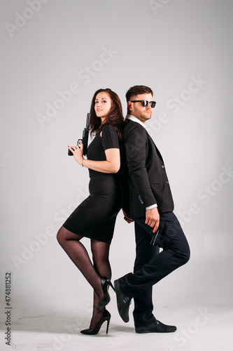young couple, in black suits with pistols in their hands on a white background in the studio, they depict security guards, bodyguards or agents.