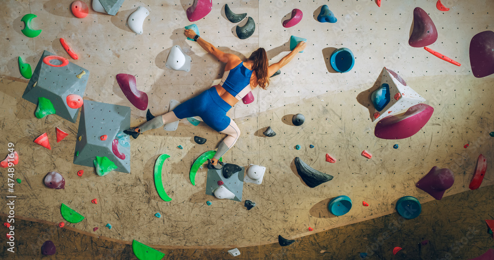 Athletic Female Rock Climber Practicing Solo Climbing on Bouldering Wall in a Gym. Female Exercising at Indoor Fitness Facility, Doing Extreme Sport for Her Healthy Lifestyle Training. Shot from Back. Athletic Female Rock Climber Practicing Solo Climbing on Bouldering Wall in a Gym. Female Exercising at Indoor Fitness Facility, Doing Extreme Sport for Her Healthy Lifestyle Training. Shot from Back.