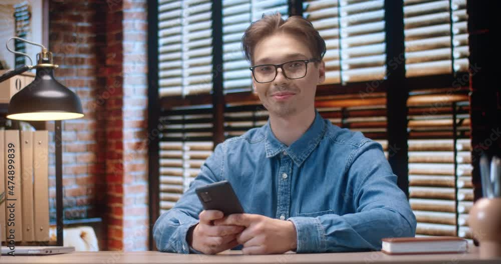 Portrait of young smart Caucasian guy with glasses sitting in cozy home office holding smartphone in hands and smiling at camera. Portrait of young smart Caucasian guy with glasses sitting in cozy home office holding smartphone in hands and smiling at camera.