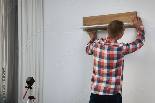 Obraz na plátně Man using cross line laser level for hanging wooden shelf on light wall indoors,