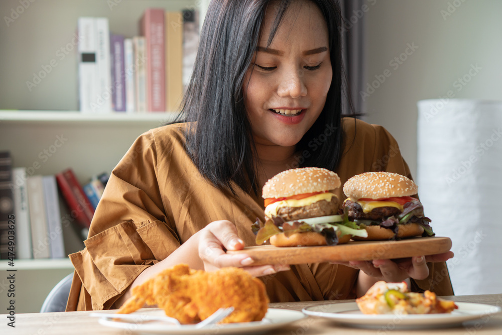 Hungry overweight woman holding hamburger on a wooden plate, Fried Chicken and Pizza on table, During work from home, gain weight problem. Concept of binge eating disorder (BED). Hungry overweight woman holding hamburger on a wooden plate, Fried Chicken and Pizza on table, During work from home, gain weight problem. Concept of binge eating disorder (BED).