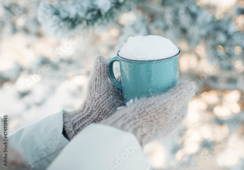 Winter image of woman's hands in the wool mittens holding vintage blue mug full of clear white snow. Winter freshness concept. Awesome snowy forest in sunny winter morning. Winter weekend out of town.
