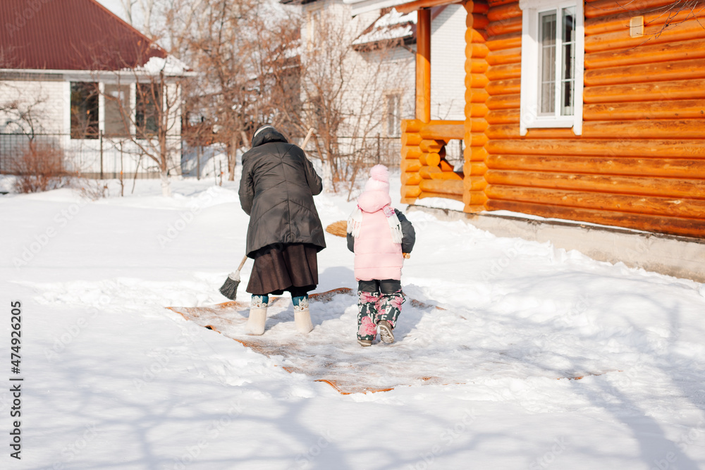Woman teaches child to clean carpet. Family is engaged in ecological carpet cleaning by traditional method with help of fresh snow and broom near wooden cottage in winter, rear view.