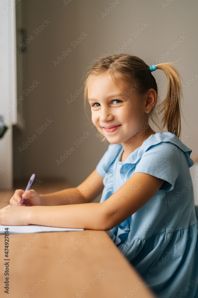 Vertical portrait of cherrful pretty primary girl child drawing sitting at home table by window looking at camera. Adorable little preschool kid studying alone making note with pen at bedroom Vertical portrait of cherrful pretty primary girl child drawing sitting at home table by window looking at camera. Adorable little preschool kid studying alone making note with pen at bedroom
