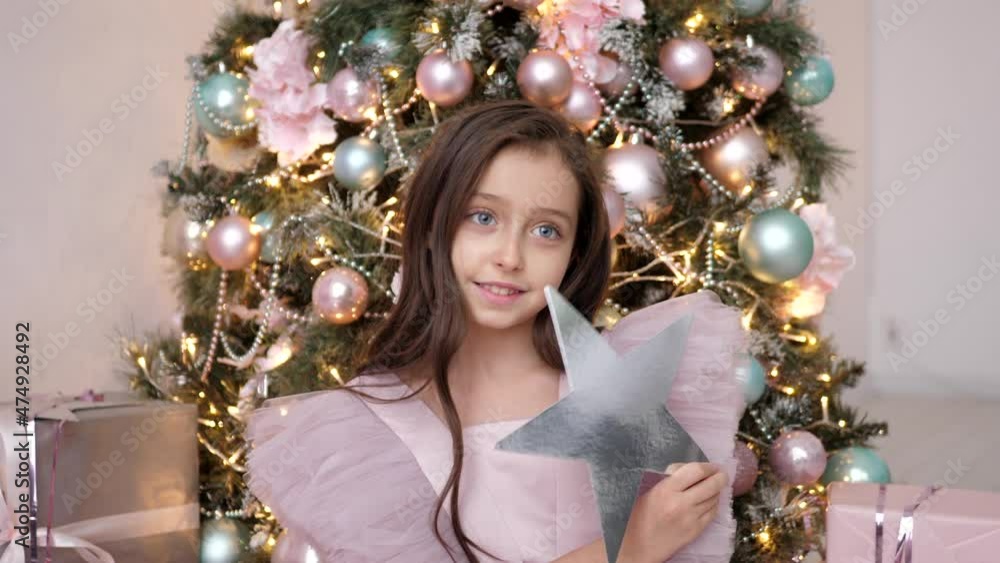 beautiful Caucasian girl judges at the Christmas tree in a pink dress and holds a star beautiful Caucasian girl judges at the Christmas tree in a pink dress and holds a star