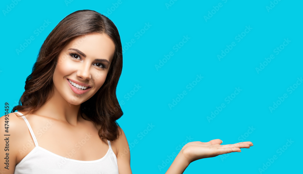 Beautiful woman in white casual wear holding hand with flat palm, giving, advertising some product showing copy space free area. Aqua blue green color background. Brunette girl at studio.