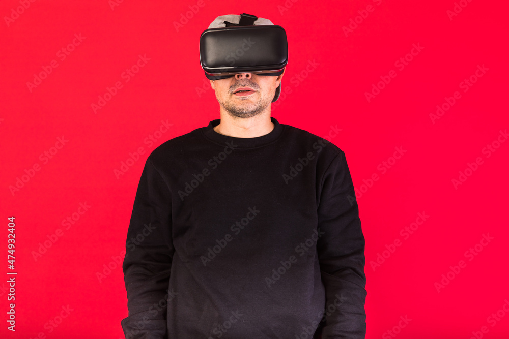 Young man with short beard, wearing black sweatshirt, cap and virtual reality glasses amazed, looking straight ahead, on red background. Technology, VR, computing and hobbies concept. Young man with short beard, wearing black sweatshirt, cap and virtual reality glasses amazed, looking straight ahead, on red background. Technology, VR, computing and hobbies concept.