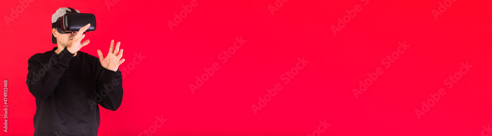 Banner of young man with short beard, wearing black sweatshirt, cap and virtual reality glasses amazed, touching something virtually, on red background. Technology, VR, computing and hobbies concept. Banner of young man with short beard, wearing black sweatshirt, cap and virtual reality glasses amazed, touching something virtually, on red background. Technology, VR, computing and hobbies concept.