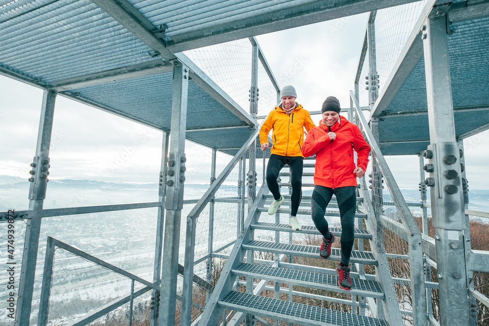 Two cheerfully smiling bright sporty clothes dressed men running down by huge steel industrial stairs with picturesque winter city landscape view. Men's friendship and healthy lifestyle concept photo.