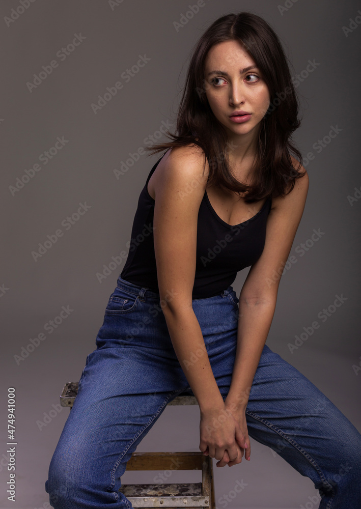 Fashion model sit on chair in a blu jeans and black undershirt barefoot on studio gray background Fashion model sit on chair in a blu jeans and black undershirt barefoot on studio gray background