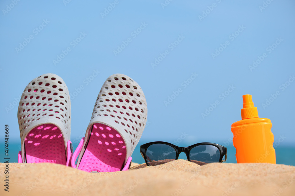 Closeup of clogs shoes, sunscreen and black protective sunglasses on sandy beach at tropical seaside on warm sunny day. Summer vacation concept