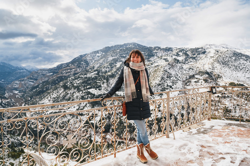 Young woman standing at mountain top observation deck over Vouraikos gorge. Mega Spileon Monastery terrace with splendid snow-covered mountains view. Winter vacation. Greece. photo