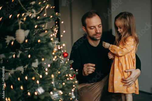 Little girl in yellow dress decorates christmas tree at home with dad