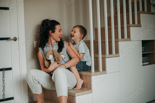 A young girl of 30 years old brunette with long hair in a white shirt with blue stripes and white jeans fooling around at home with her blond son in a blue shirt and blue shorts