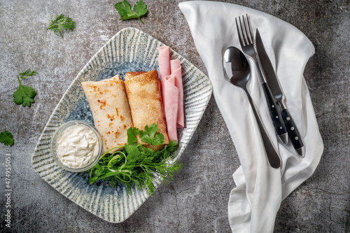 Pancakes filled with ham in a plate with parsley and dill greens and sour cream on a gray stone table photo