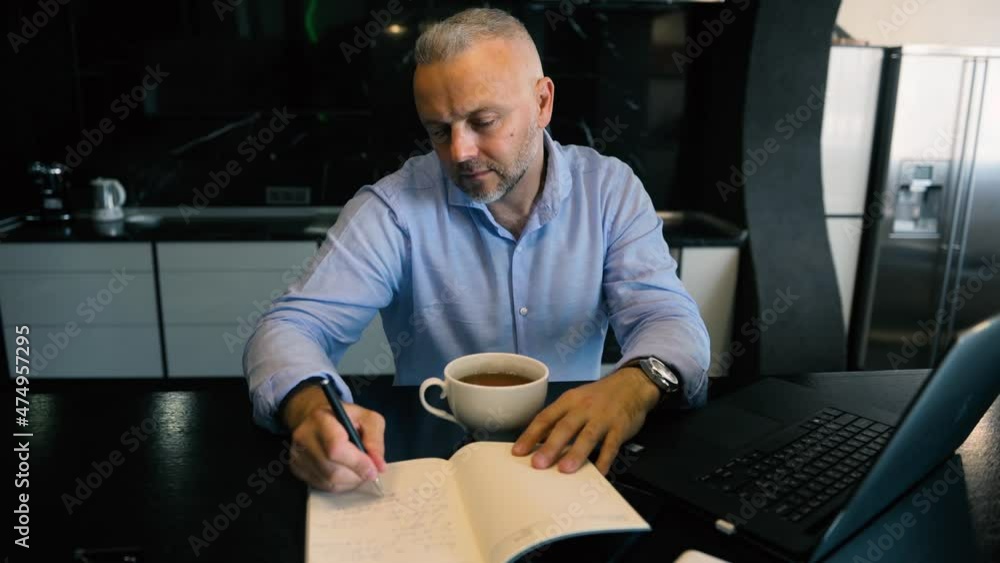 Handsome middle aged man, successful businessman entrepreneur writing on notepad, making notes while enjoying morning coffee, sitting t kitchen table in front of a laptop Handsome middle aged man, successful businessman entrepreneur writing on notepad, making notes while enjoying morning coffee, sitting t kitchen table in front of a laptop