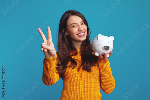 Excited young girl in casual orange sweater holding white piggy bank with lots of money, showing peace gesture over blue background photo