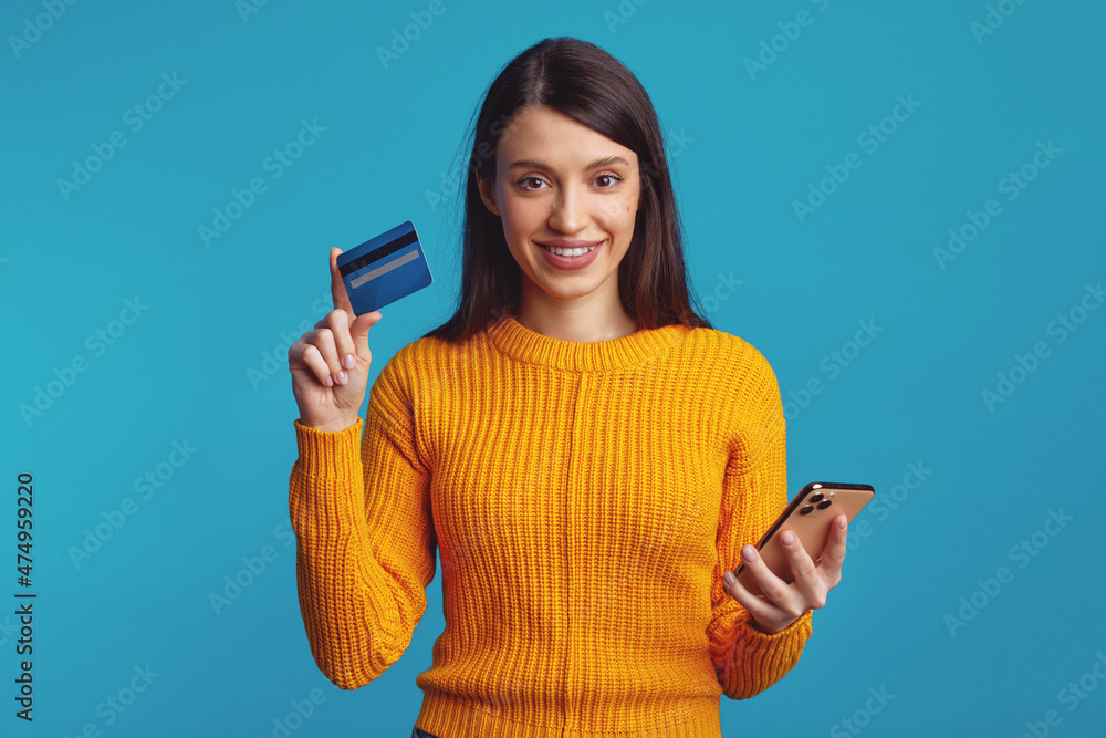 Portrait of a happy young brunette girl showing plastic credit card while holding mobile phone isolated over blue background Portrait of a happy young brunette girl showing plastic credit card while holding mobile phone isolated over blue background