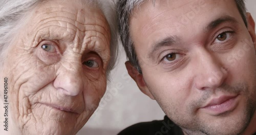 The face of a grandmother and an adult son. Portrait of an old woman with deep wrinkles and a young man sitting together on the sofa at home.
