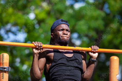 Close up of a slim black guy in a cap doing pulls up on a crossbar during workout in the park