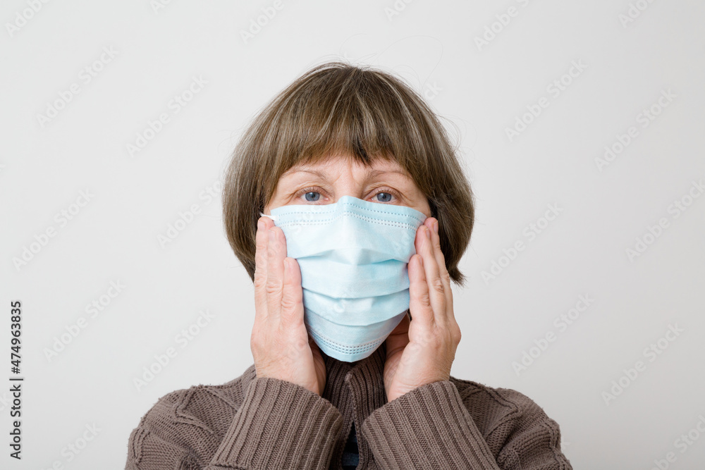 Old mature woman hands touching respirator mask on face for mouth and nose covering. Isolated on light gray background. Head closeup. First protection in virus time. Healthcare concept. Front view. Old mature woman hands touching respirator mask on face for mouth and nose covering. Isolated on light gray background. Head closeup. First protection in virus time. Healthcare concept. Front view.