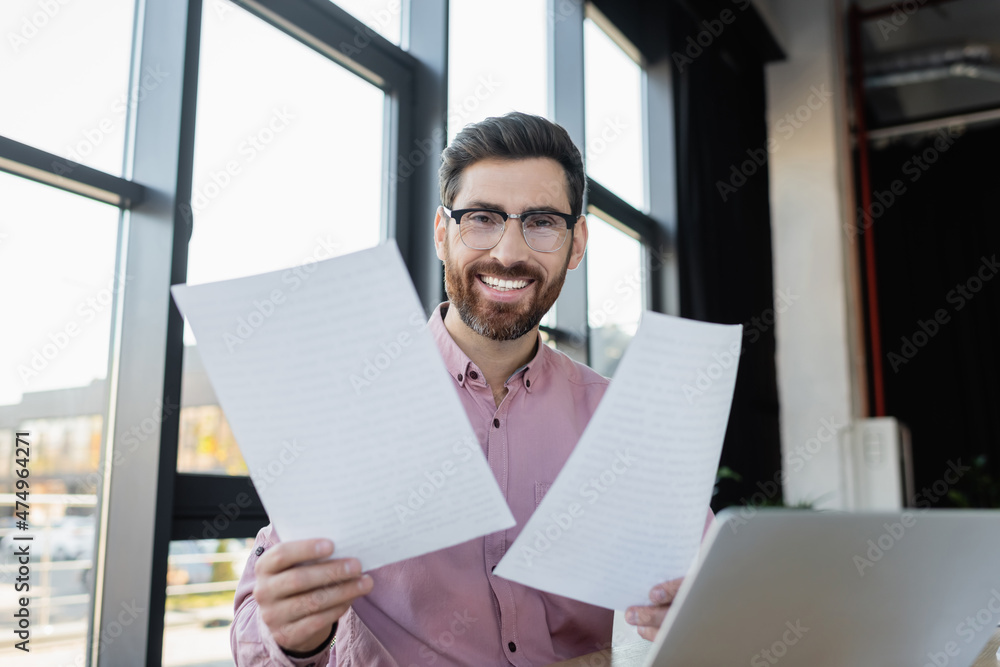 Smiling businessman holding documents near laptop in office. Smiling businessman holding documents near laptop in office.