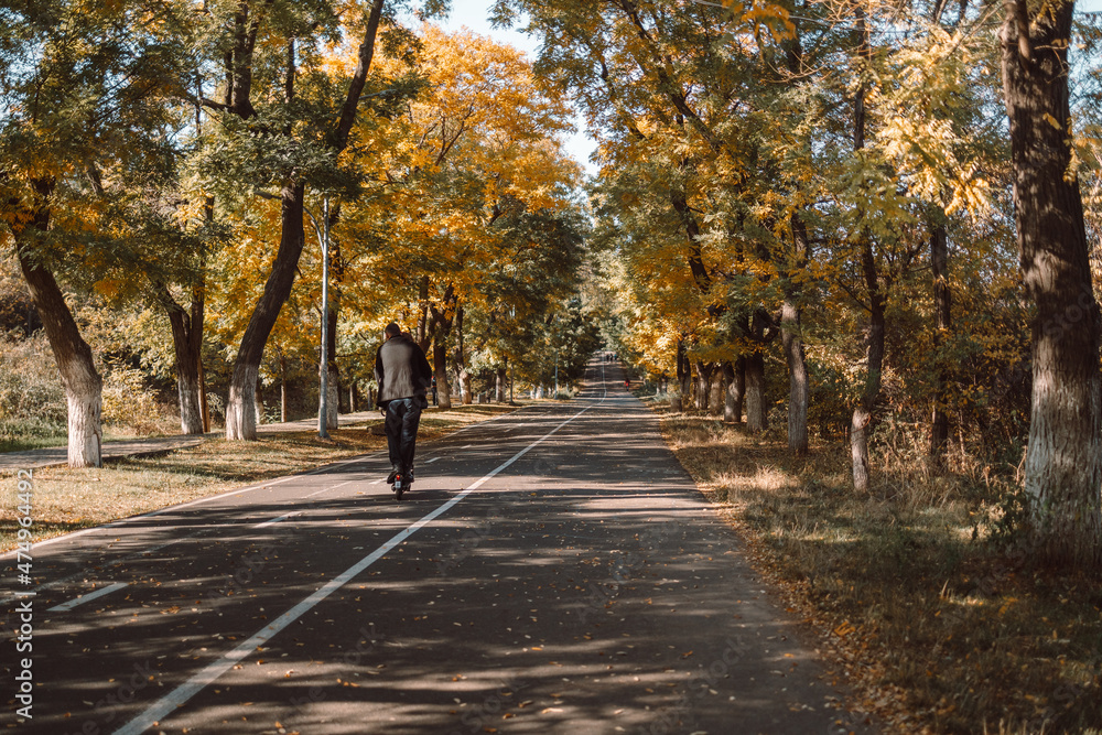 Guy rides an electric scooter on a rural park road. Autumn landscape. Fall scene.Trees and leaves in sunlight rays Guy rides an electric scooter on a rural park road. Autumn landscape. Fall scene.Trees and leaves in sunlight rays