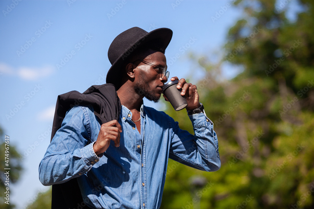 Young confident black man in a hat walking in a summer park holding a jacket over his shoulder and drinking coffee Young confident black man in a hat walking in a summer park holding a jacket over his shoulder and drinking coffee