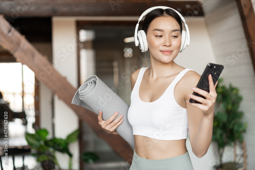 Young asian woman standing at home in headphones, listening music and going workout on floor mat, practice yoga or meditate in living room photo