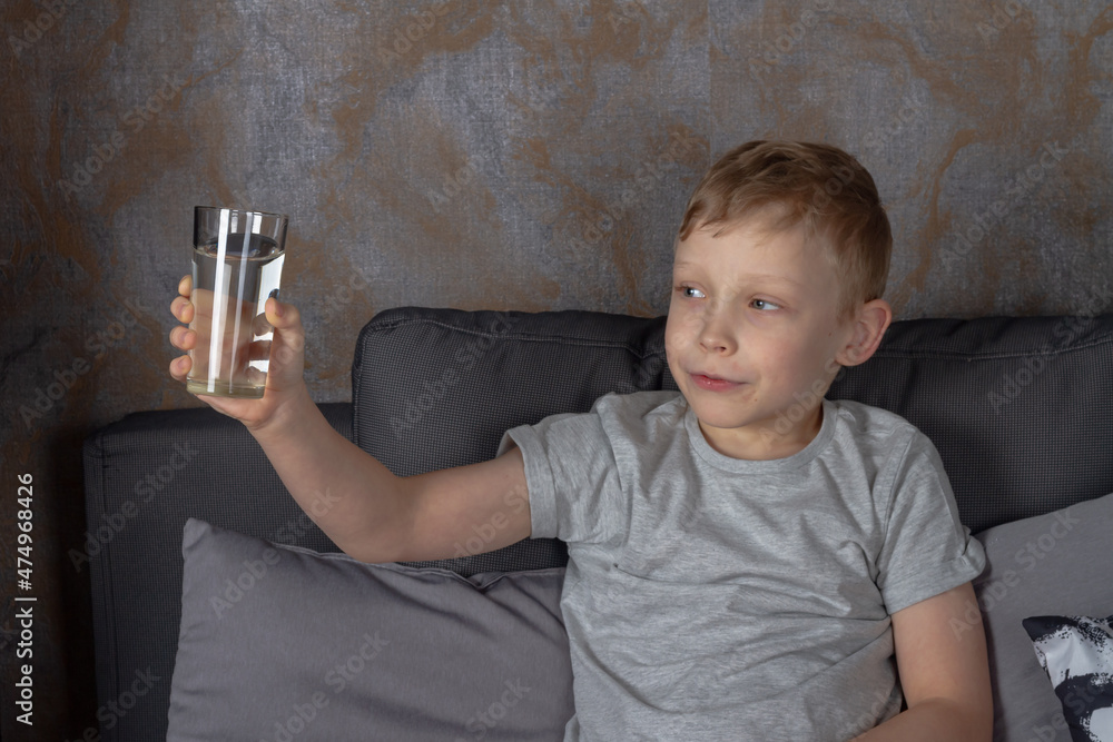 Naklejka premium A caucasian boy drinks water from a glass while sitting on a gray sofa