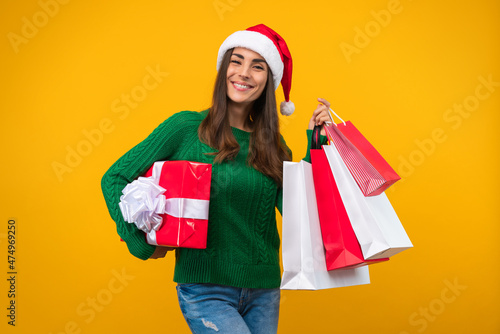 Studio shot of attractive smiling young woman in Santa hat posing with gift box and a bunch shopping bags in hands. Isolated over yellow background