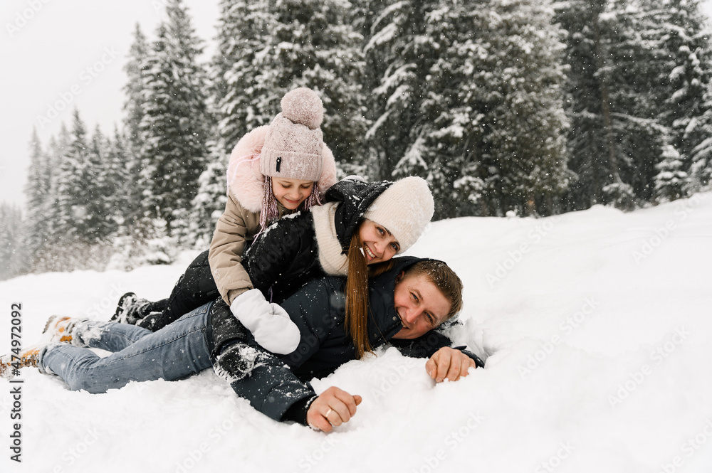 Obraz premium Happy family have fun in winter forest. Mother, father and dauther lie on the ground and playing with snow. Family concept. Enjoying spending time together