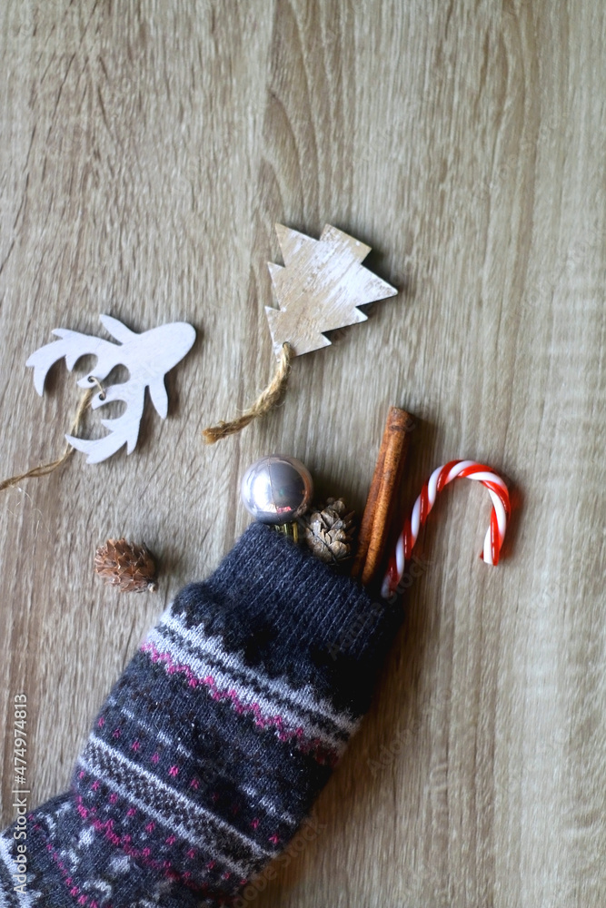 Fototapeta premium Wool stocking filled with small Christmas ornaments, cinnamon stick and candy cane. Wooden background, flat lay.