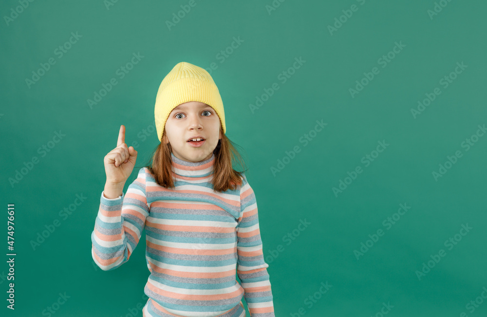 a beautiful little boy with long hair in a yellow hat stands thoughtfully raising his finger to the top on an isolated green background. a beautiful little boy with long hair in a yellow hat stands thoughtfully raising his finger to the top on an isolated green background.