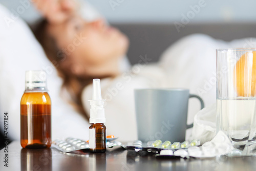 Sick young woman covered with a blanket lying in bed with high fever and a flu. A lot of medicines on table in bedroom
