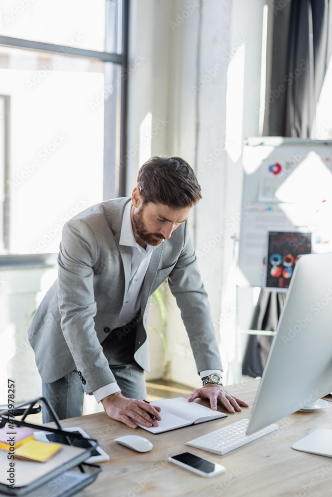 Businessman looking at notebook near devices and papers in office. Businessman looking at notebook near devices and papers in office.