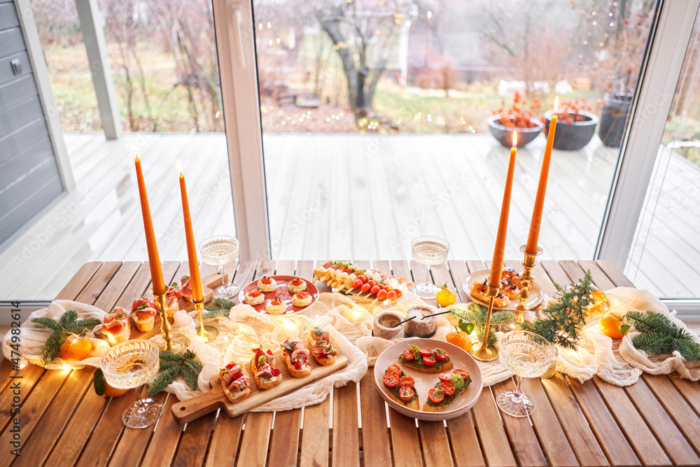 Christmas dinner feast. A small table is served with snacks, bruschettas, and canapes. A decorated dining table with champagne glasses, candles and christmas tree an garland in background
